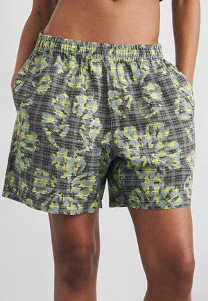 Mann iført knelang rutete shorts med gult og blått blomsterprint og elastisk midje, stående med hendene i lomma.