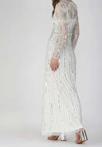 Lungo vestito bianco con paillettes argentate disposte in strisce verticali, maniche lunghe trasparenti e un corpetto decorato con perline e dettagli floreali. Tacchi argento.
