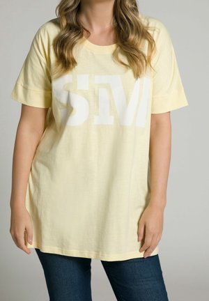 Kobieta w jasnofioletowej, luźnej koszulce typu oversize z białymi, częściowo widocznymi literami "SIV" oraz ciemnoniebieskich dżinsach, stojąca na jednolitym tle.