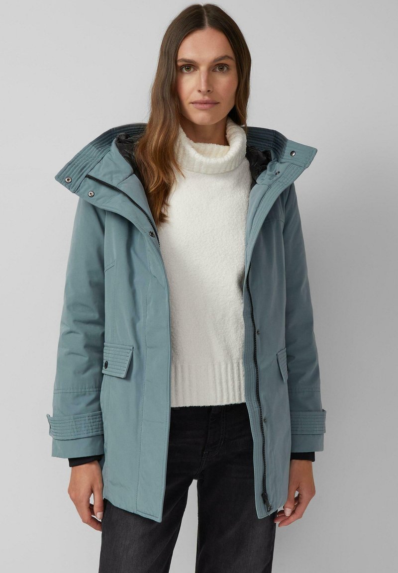 Svijetloplava parka s kapuljačom, izrađena od glatkog, vodoodpornog materijala. Ima zatvaranje na zip, duge gumbe i podstavljene rukave. Nosena preko bijelog džempera.