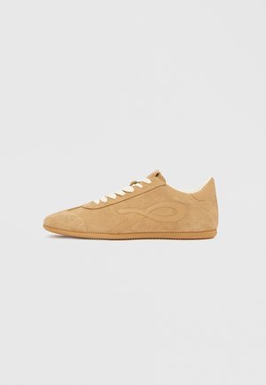 Beige Wildledersneaker mit abgerundeter Spitze, kontrastierenden Schnürsenkeln und einem strukturierten Obermaterial mit einem markanten Design an der Seite.