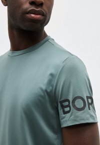 Άνδρας που φοράει ένα χαμηλόχρωμο τιρκουάζ κοντομάνικο πουκάμισο με μαύρα υφασμένα γράμματα "BOR" ορατά στο δεξί μανίκι, μπροστά σε απλό φόντο.