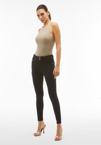 Monos de color beige con cuello alto, combinados con jeans ajustados negros que cuentan con dos bolsillos frontales y botones plateados. Sandalias con tacón transparente.