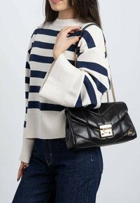 Bolso de cuero negro acolchado con un cierre metálico dorado, llevado con una cadena al hombro. Modelo con suéter de rayas y vaqueros azules.