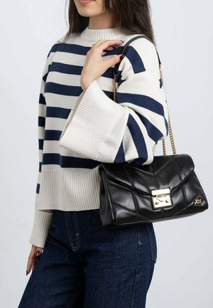 Bolso de cuero negro acolchado con un cierre metálico dorado, llevado con una cadena al hombro. Modelo con suéter de rayas y vaqueros azules.