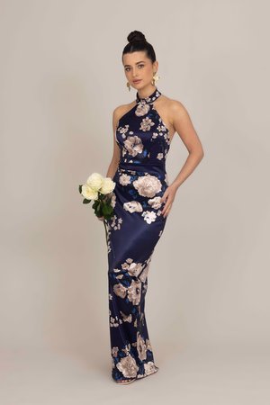 Floor-length navy satin dress with floral patterns in pink and blue, featuring a halter neck and a fitted silhouette, paired with white roses.