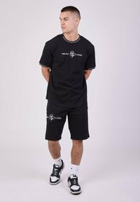 Conjunto de camiseta de manga corta negra y pantalones cortos, con texto de la marca en blanco y acentos de tachuelas en el cuello y los puños. Combinado con zapatillas negras.