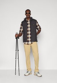 Gray vest over a plaid shirt, beige pants, and gray sneakers. Holding a black trekking pole. Accessories include white sunglasses.