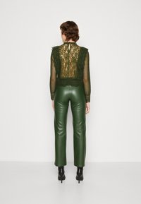Blusa verde in pizzo con scollatura a forma di onde e maniche lunghe, abbinata a pantaloni in pelle verde aderenti, vista da dietro. Tacchi neri.