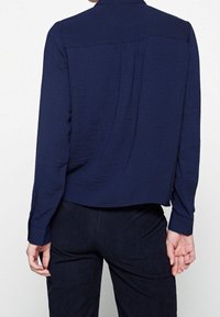 Blusa blu navy a maniche lunghe con tessuto strutturato, retro plissettato e polsini con bottone, dotata di colletto sagomato e vestibilità morbida.