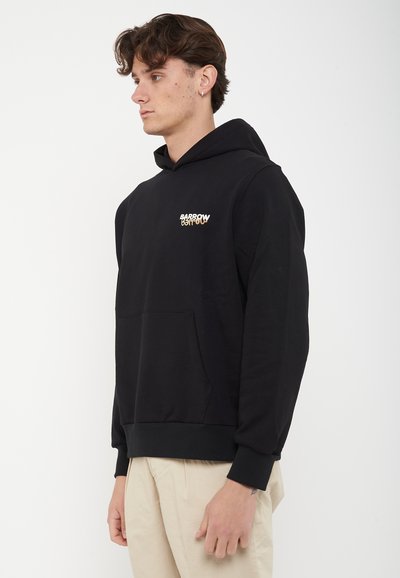 Νέος άνδρας με μαύρο φούτερ με κουκούλα και το λογότυπο "Barrow" στο στήθος, στέκεται πλαγίως μπροστά σε απλό λευκό φόντο.