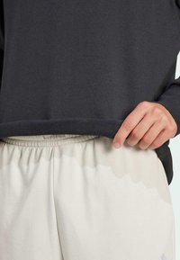 Mørkegrå sweatshirt med en struktureret kant, parret med lyse beige bukser med elastisk talje. Nærbillede af en hånd, der hviler på sweatshirten.