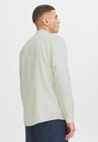 Camisa de manga larga en color beige claro, confeccionada con tela texturizada, con un collar puntiagudo y puños abotonados, vista desde la parte trasera.