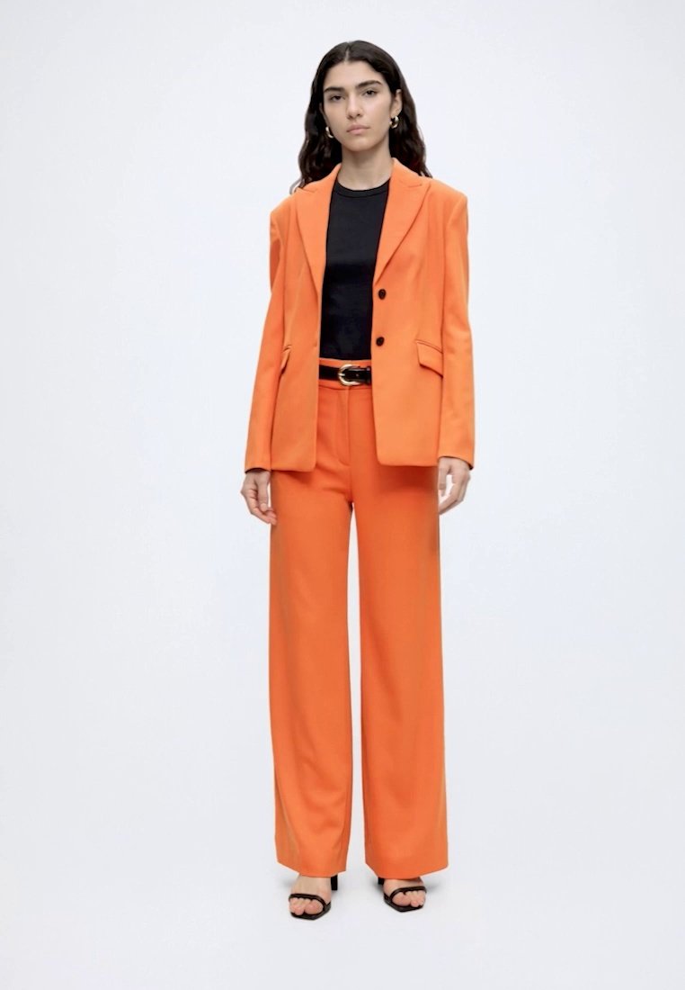 Femme debout, face à l'avant, portant un blazer et un pantalon orange, un haut noir et une ceinture, et des sandales à talons noires, sur fond uni.