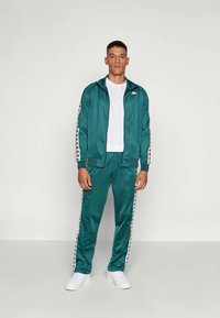 Herre teal treningsdrakt i glatt stoff; zip-up jakke med svarte og hvite sidestiper; matchende joggebukser med elastiske mansjetter; kombinert med hvite sko.