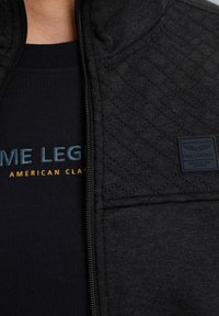 Gros plan sur une veste noire matelassée avec fermeture éclair à côté d'une chemise bleu foncé présentant un texte jaune et bleu partiellement visible.