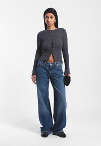 Szara, prążkowana bluzka z długim rękawem, zapinana na guziki, o skróconym fasonie, w połączeniu z luźnymi niebieskimi dżinsami. Czarna torebka kopertówka, stojąca na białym tle.