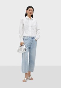 Chemisier blanc avec des détails en pli, jeans en denim bleu clair à jambes larges, sac à main métallique argenté et sandales métalliques. Fond neutre.