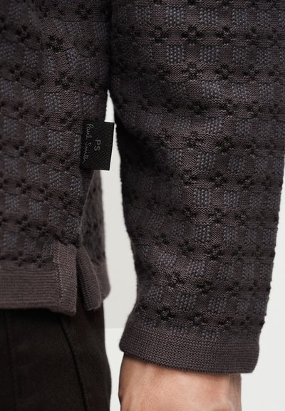 Μανίκι πουλόβερ από σκούρο γκρι με πλεκτή υφή και μια μικρή μαύρη υφασμάτινη ετικέτα με την ονομασία "PS Paul Smith" κοντά στην μανσέτα.
