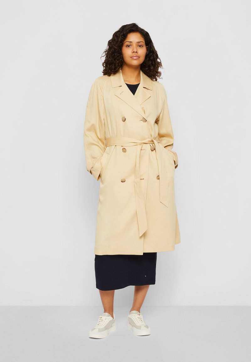 Trench coat doppiopetto beige con cintura, maniche lunghe e bottoni abbinati. Indossato sopra una gonna blu scuro e sneakers.
