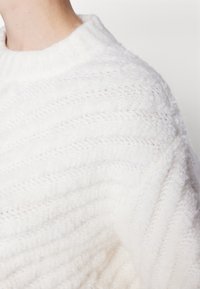 Maglione bianco lavorato a maglia con colletto a coste e un motivo ondulato e texturizzato. Il tessuto appare morbido e peloso con una leggera trasparenza.