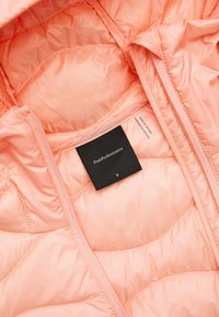 Interior de chaqueta acolchada color durazno con textura suave. Presenta una cremallera, forro y una etiqueta negra que dice "PeakPerformance" en tamaño S.