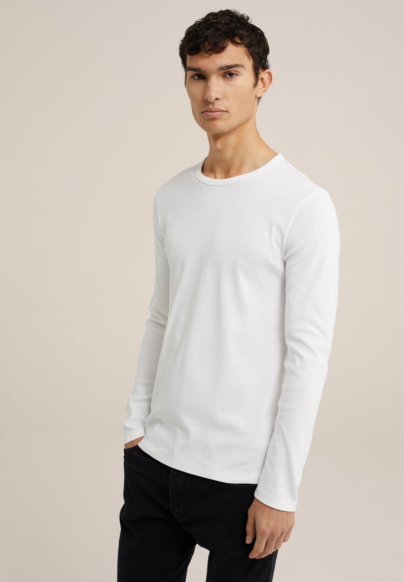 Camiseta blanca de manga larga, con un diseño ajustado y cuello redondo. La tela parece suave y lisa, combinada con jeans oscuros.
