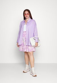 Camisa de botão oversized lilás claro, top crop branco, saia em tie-dye pastel, mala branca e ténis altos brancos com solas castanhas.
