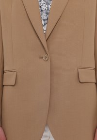 Beige bleiseri, jossa on yksi nappi ja kaksi etutaskua. Muotoonommeltu malli, sileä pinta ja hienovarainen tikkausdetail.