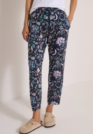 Personne portant un pantalon à cordon bleu marine à imprimé floral et des chaussures beige à enfiler, debout avec une main dans la poche.