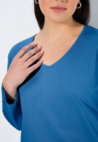 Blaues V-Ausschnitt-T-Shirt mit langen Ärmeln, aus weichem Stoff, mit sauberem Ausschnitt und subtiler Textur. Hand ruht auf dem Brustbereich.