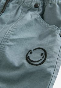 Tissu bleu clair avec un visage souriant brodé en noir sur la poche, avec une texture en grille subtile et des coutures visibles ainsi que des boutons-pression.