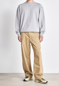 Felpa grigia con collo a giro e polsini a coste, abbinata a pantaloni beige a gamba larga. La calzatura include sneakers con dettagli neri e marroni.