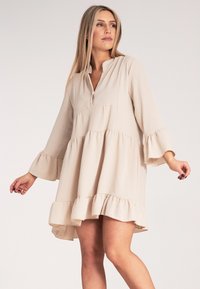 Beżowa sukienka z długim rękawem, dekoltem w kształcie V, falbaną u dołu i warstwowym designem. Wykonana z teksturowanej tkaniny, o swobodnym kroju i luźnej sylwetce.