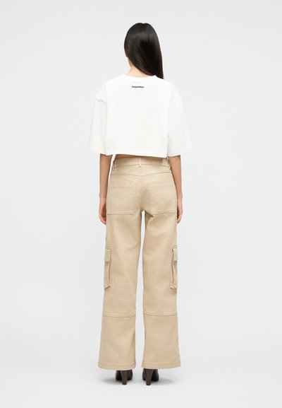 Γυναίκα με μακριά σκούρα μαλλιά, φοράει λευκό κοντό μπλουζάκι και μπεζ φαρδιά παντελόνια cargo, στέκεται μπροστά σε ένα απλό λευκό φόντο.