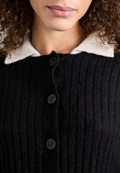 Μαύρο καρό ζακέτα με κρεμ γιακά, με πέντε μαύρα κουμπιά μπροστά, επιδεικνύοντας μια απαλή, υφή υφάσματος.