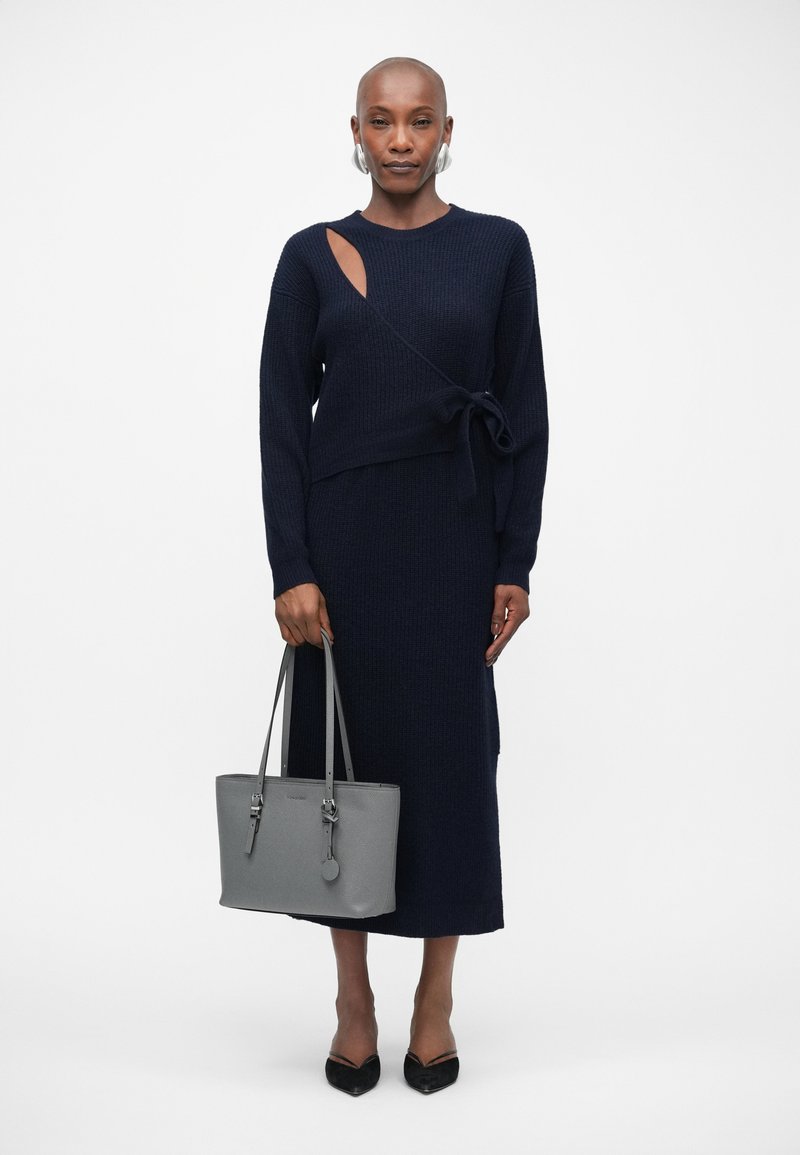 Femme portant une robe en tricot bleu marine avec une découpe sur le côté et un lien, tenant un tote gris, et portant des talons noirs pointus sur un fond uni.