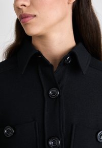 Camisa negra de botones con un cuello estructurado, que presenta grandes botones negros brillantes y dos bolsillos en el pecho, hecha de una tela texturizada.