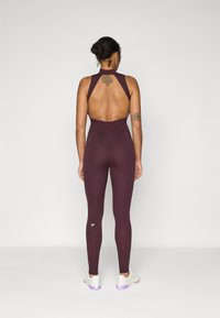 Maroon athletic bodysuit with a high back, cut-out details, and fitted leggings. Features a small logo on the lower leg. Smooth texture.