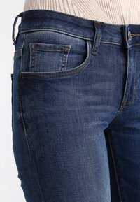 Jeans de mezclilla azul oscuro con un diseño clásico de cinco bolsillos, un ligero desgaste y un cierre de botón en la cintura.