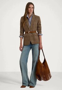 Mulher a usar um blazer xadrez castanho cingido na cintura, camisa riscada azul, jeans de perna larga, sapatos castanhos de bico fino e a segurar uma mala de franjas castanha.