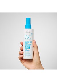 Spray après-shampoing BC Moisture Kick dans un flacon blanc avec un bouchon-pompe bleu, présentant un texte et des icônes bleus audacieux, tenu par une main. Taille de 200 ml.