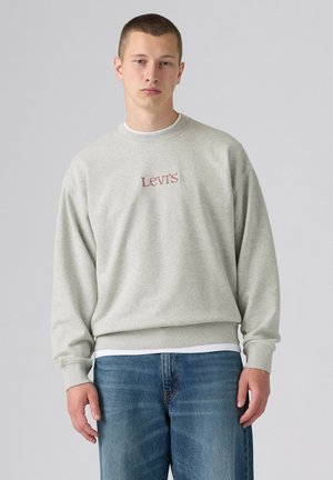 Młody mężczyzna ubrany w jasnoszarą bluzę Levi's i niebieskie jeansy, stojący na tle jednolitego jasnoszarego tła.