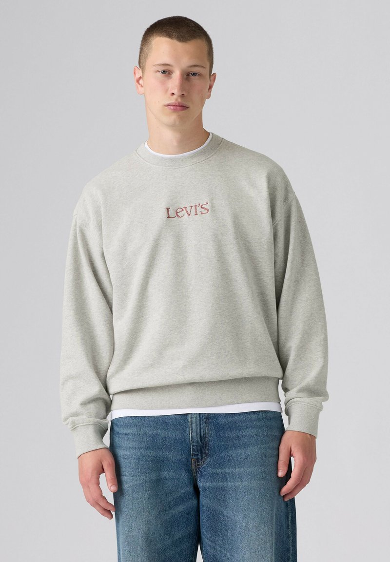 Giovane uomo che indossa una felpa Levi's grigio chiaro e jeans blu, in piedi contro uno sfondo grigio chiaro uniforme.