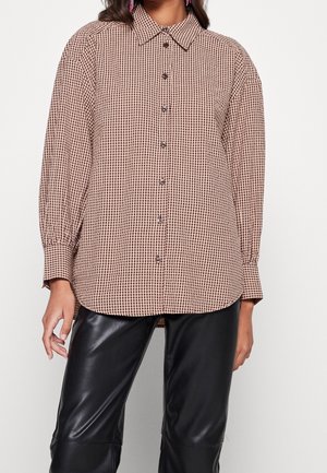 Vrouw met een bruin-zwart geruit overhemd met lange mouwen en een zwarte leren broek, staand tegen een witte achtergrond.