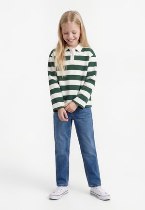 Naeratav tüdruk, kellel on pikad blondid juuksed, kannab rohelist ja valget triibulist pika varrukaga polo, siniseid teksapükse ja valgeid tossusid, seisab käed kokkusobitatud.