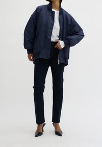 Granatowa kurtka bombowiec z zamkiem z przodu, ściągaczami przy rękawach i luźnym krojem, zestawiona z ciemnymi dżinsami i eleganckimi czarnymi szpilkami.