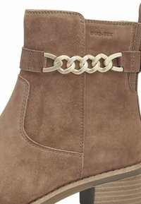 Bottine en daim marron avec un accent métallique en chaîne et un talon, matériau texturé, bout arrondi et détails de couture le long des coutures.