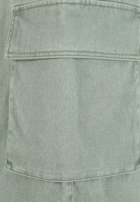 Tissu en denim vert avec un tissage texturé. Comprend une poche rectangulaire avec une couture visible le long des bords et des côtés.