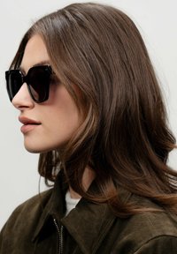 Černé oversized sluneční brýle se čtvercovým rámem, v kombinaci s tmavozelenou semišovou bundou, která má zip a vysoký límec.